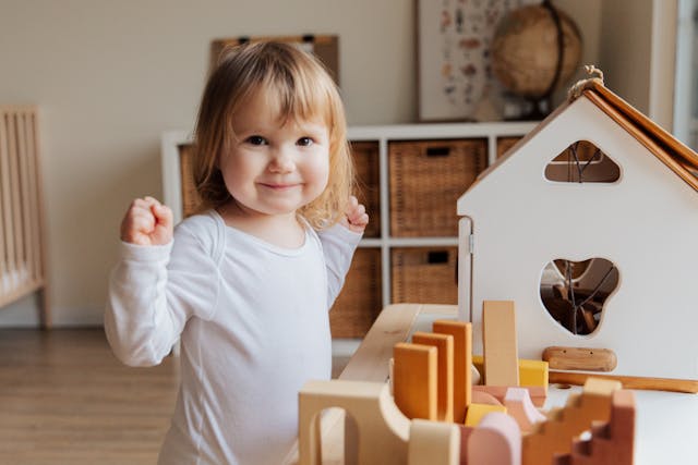 Une jeune enfant souriante joue dans une chambre lumineuse, debout à côté d'une maison de poupée en bois moderne.